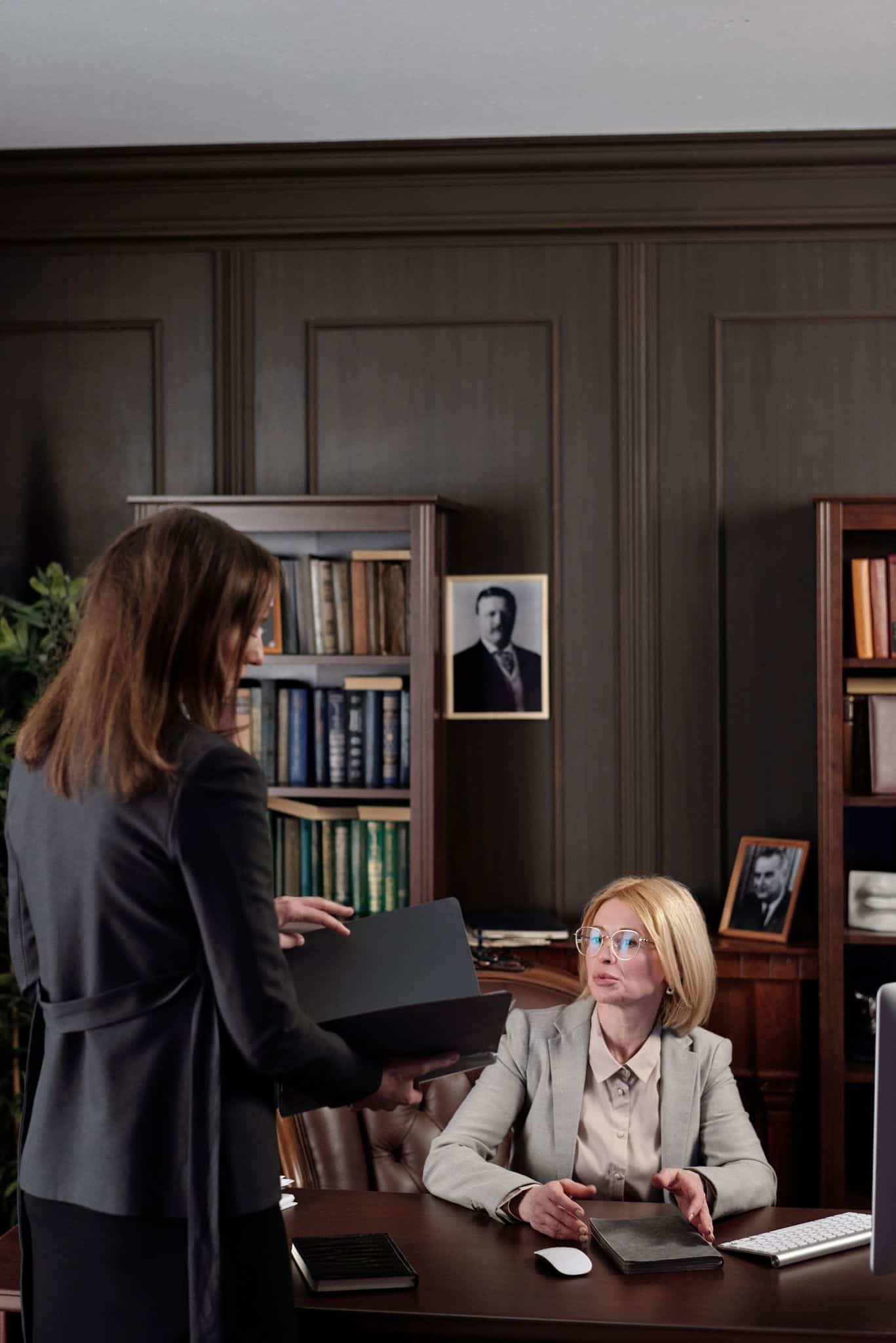 Two businesswomen at a meeting in a book-filled office. Professional and focused setting.